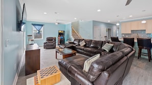 A spacious living room with a brown leather sofa, a wooden coffee table, and a chessboard. There is a TV mounted on the wall, a staircase leading to another level, and a kitchen area with wooden cabinets and a tiled backsplash.