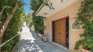 A narrow, tree-lined pathway leading to a building with a wooden door and a small potted plant beside it. - Lovely 2 Bedroom Apartment Vouliagmeni (Vari-Voula-Vouliagmeni)