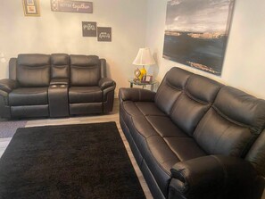 Una sala de estar con un sofá de cuero negro, un sillón reclinable de cuero negro y una mesa de centro de vidrio con una lámpara.