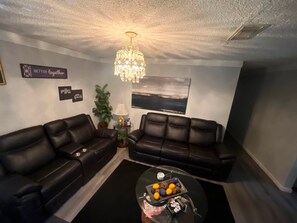 A living room with two black leather sofas, a glass coffee table with fruits, a chandelier, and wall art.