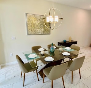 Uma sala de jantar com mesa de vidro, seis cadeiras, um lustre, um vaso decorativo e uma obra de arte na parede.