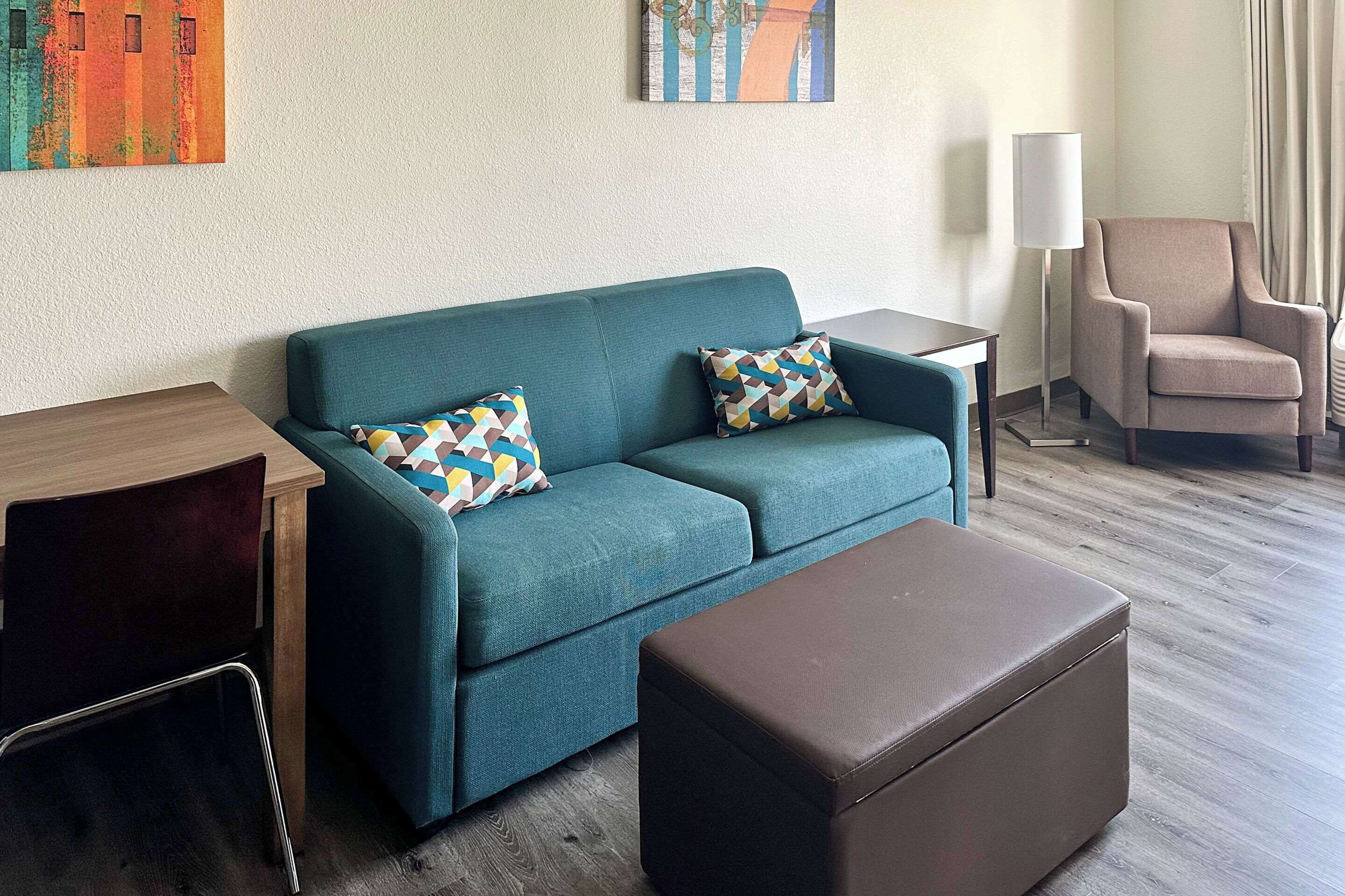 Een moderne hotelkamer met een blauwgroene bank, een bruin poefje, een houten bijzettafeltje, een klein bureau, een lamp en een stoel.