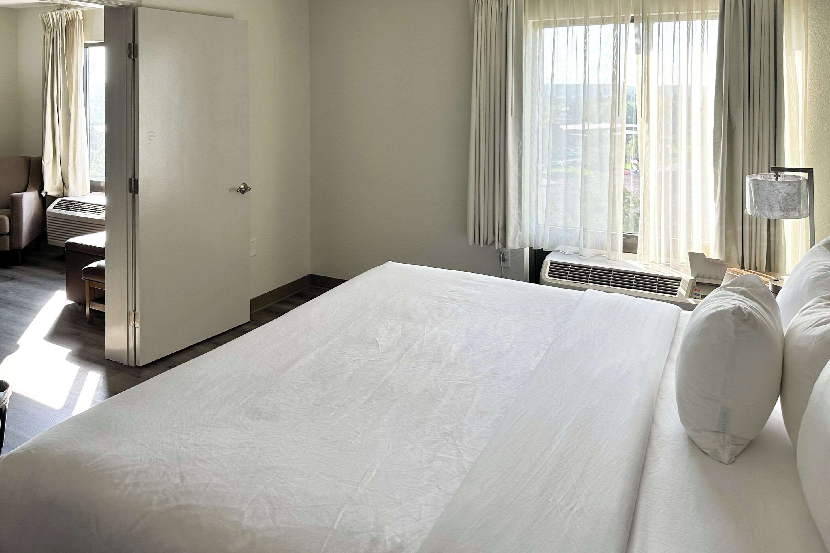 A hotel room with a large bed, a desk, and a chair. There is a window with curtains and a lamp on the desk.