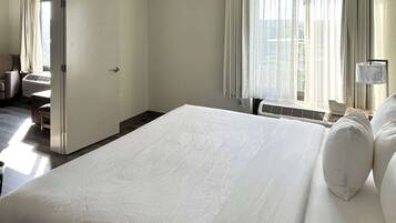 A hotel room with a large bed, a desk, and a chair. There is a window with curtains and a lamp on the desk.