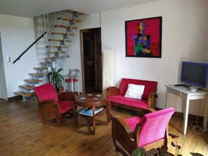 Un salon avec un escalier en bois, un canapé rouge, des chaises en bois, une petite table et un tableau accroché au mur.