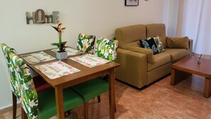 Una sala de estar con un sofá marrón, sillas estampadas en verde y flores, una mesa de centro de madera y una mesa de comedor con manteles individuales estampados.