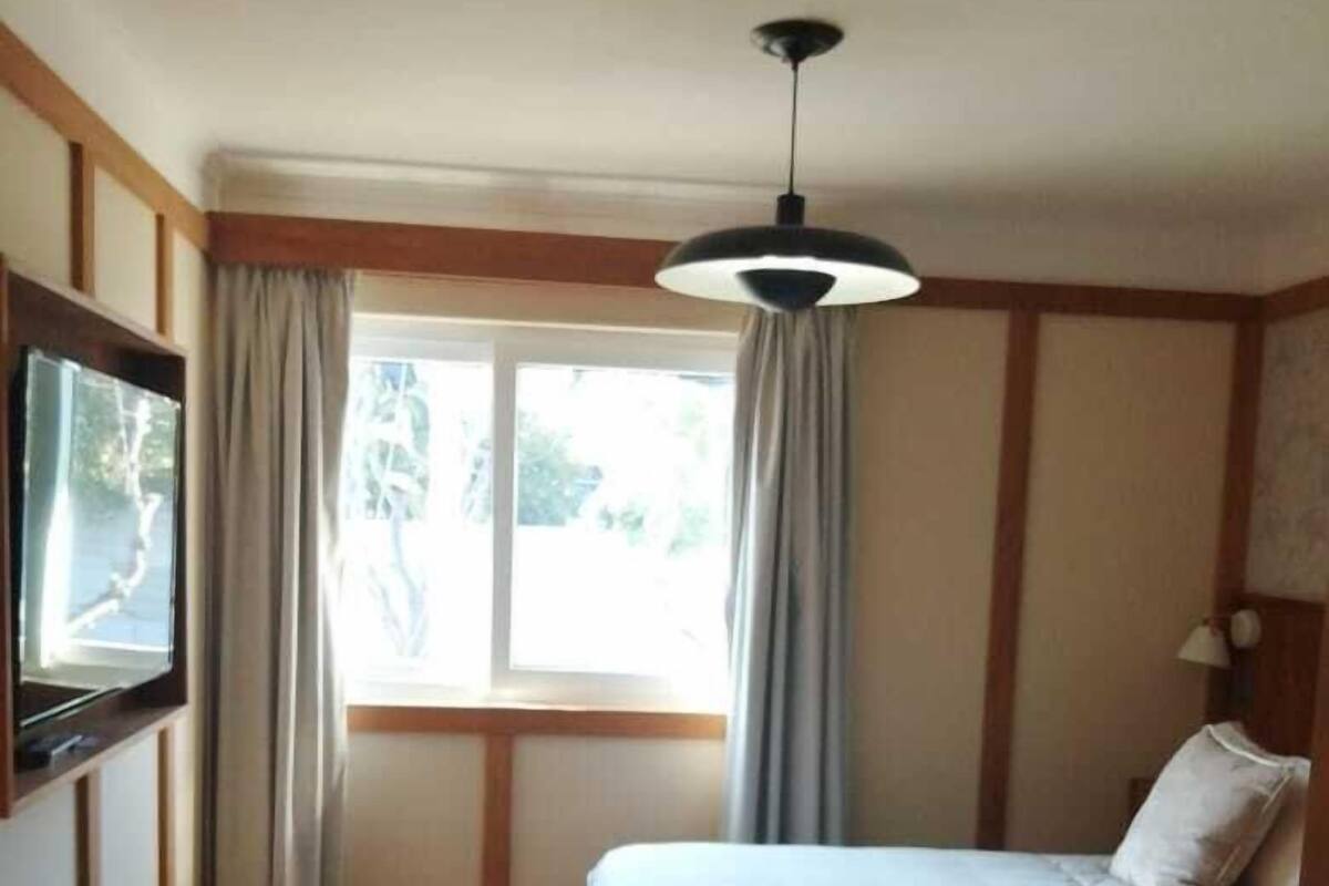 Een slaapkamer met een bed, een aan de muur bevestigde televisie, een raam met gordijnen en een plafondlamp.
