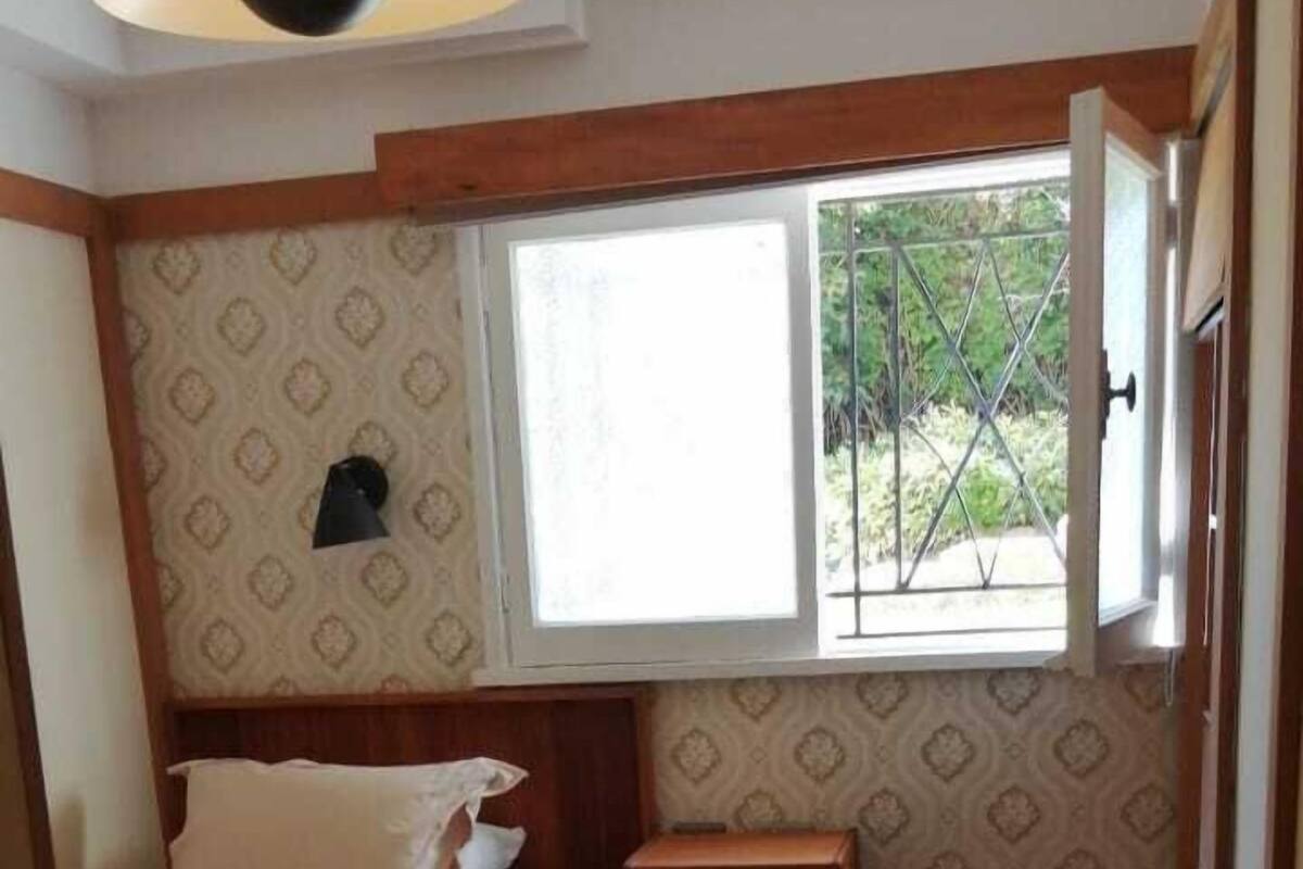 Een kleine, gezellige kamer met een bed, een nachtkastje, een raam met uitzicht op groen en behang met een patroon.