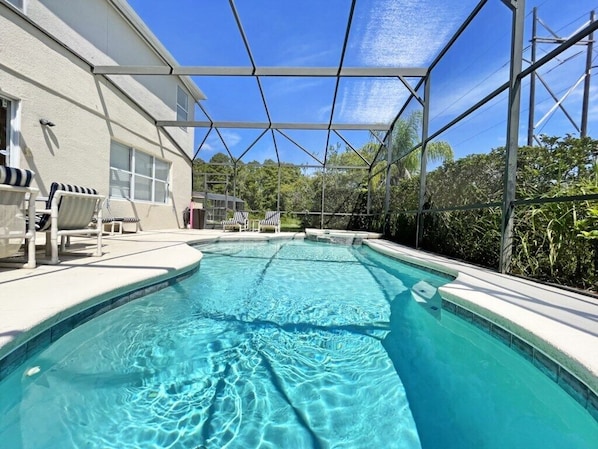 A swimming pool with a clear blue water, surrounded by a covered area with chairs and a building in the background.