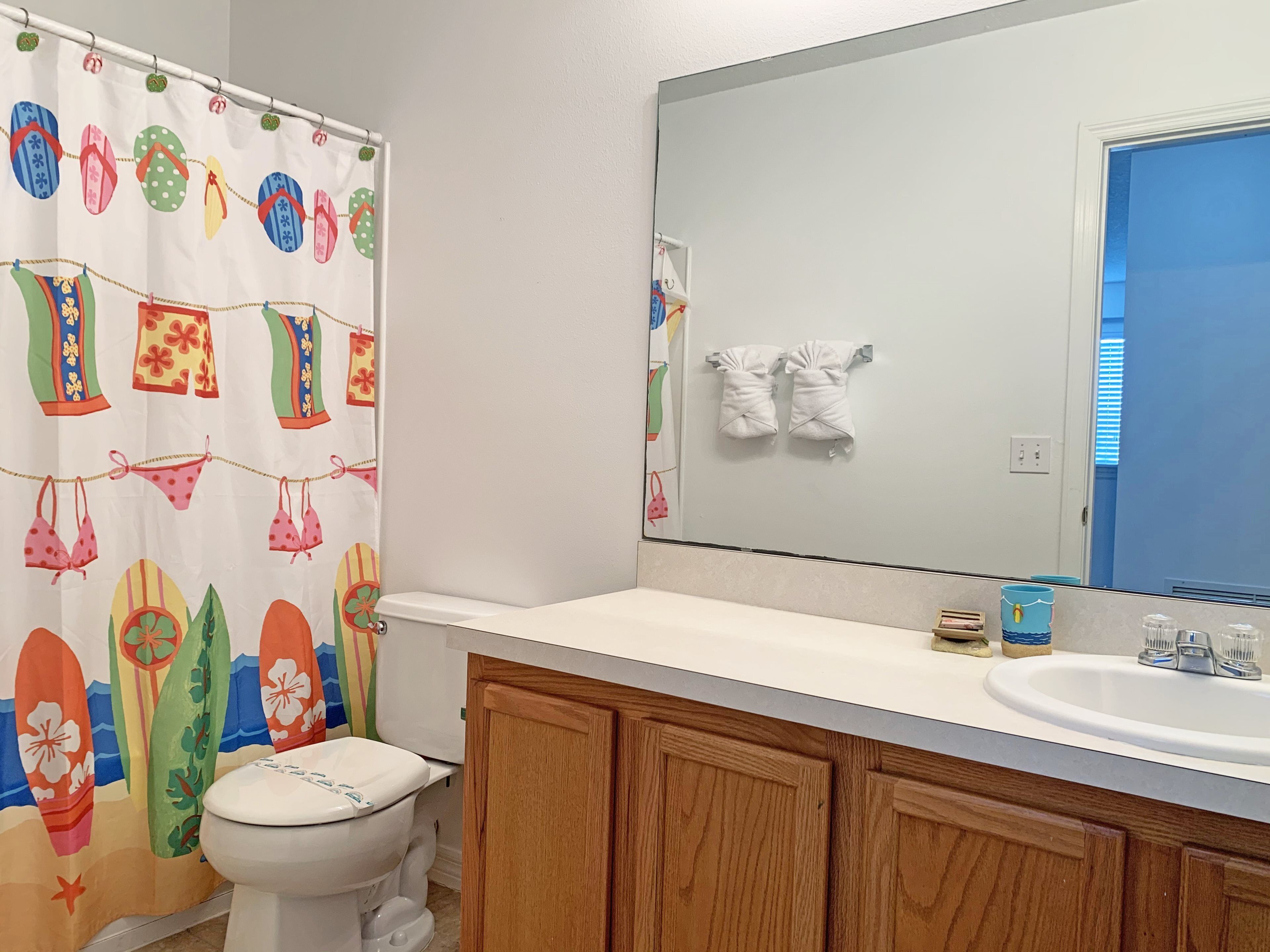 A bathroom with a shower curtain featuring a tropical design, a white toilet, wooden cabinets, a large mirror, and a sink with a blue container.