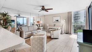 A modern living room with a sofa, armchairs, a coffee table, and a TV. There is a ceiling fan, a dining table with chairs, and a large window with a view of the outside. - ⭐Walk 2 Beach-Gulf + Pool Views-2 Pools + Hot Tub-2BR Mainsail 213⭐ (Miramar Beach)