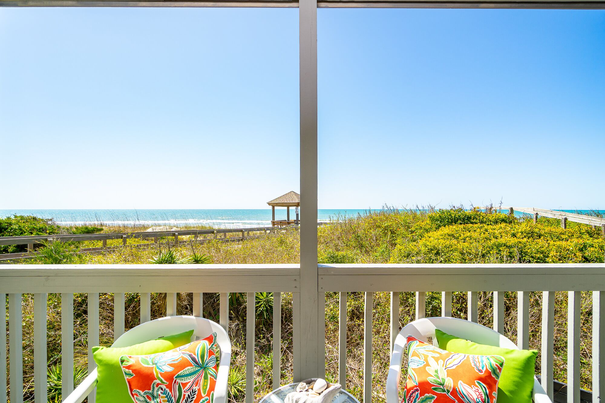 A balcony with two chairs, a view of the beach, and a gazebo in the distance.