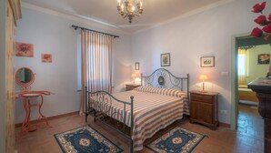 A bedroom with a bed, two bedside tables, a chandelier, a mirror, and a window with curtains. - LA ROSA CANINA     . (Castiglione del Lago)