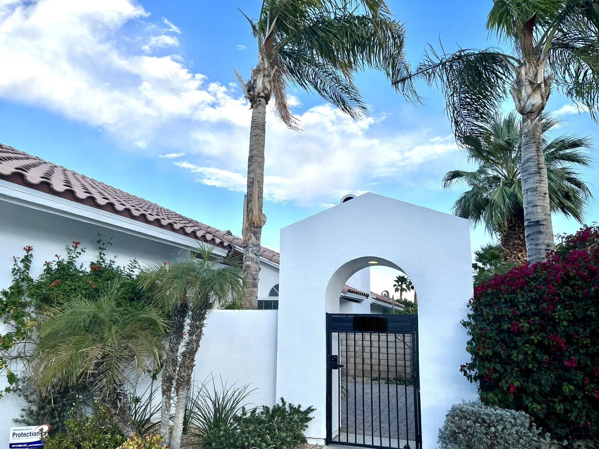 A gated entrance with a white wall and a metal gate, surrounded by palm trees and flowering plants.