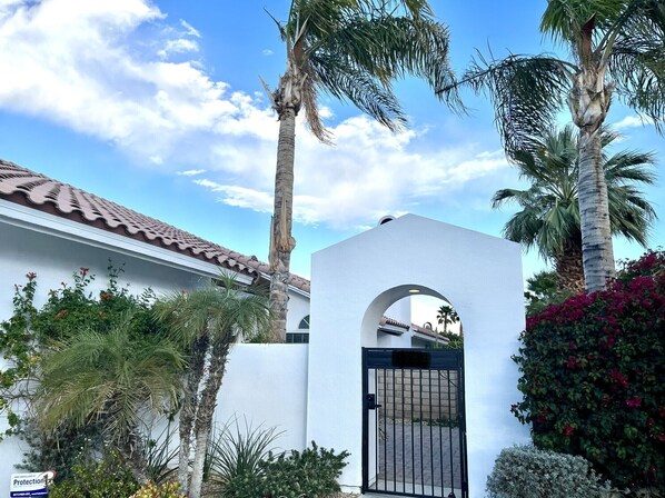 A white building with a tiled roof, a gated entrance, and palm trees.