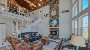 A spacious living area with a high ceiling, a stone fireplace, a large TV, and a staircase leading to an upper level.