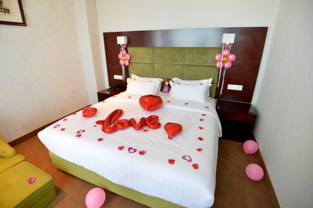 A neatly made bed with a white and green bedspread, red heart-shaped decorations spelling "love," and pink balloons.