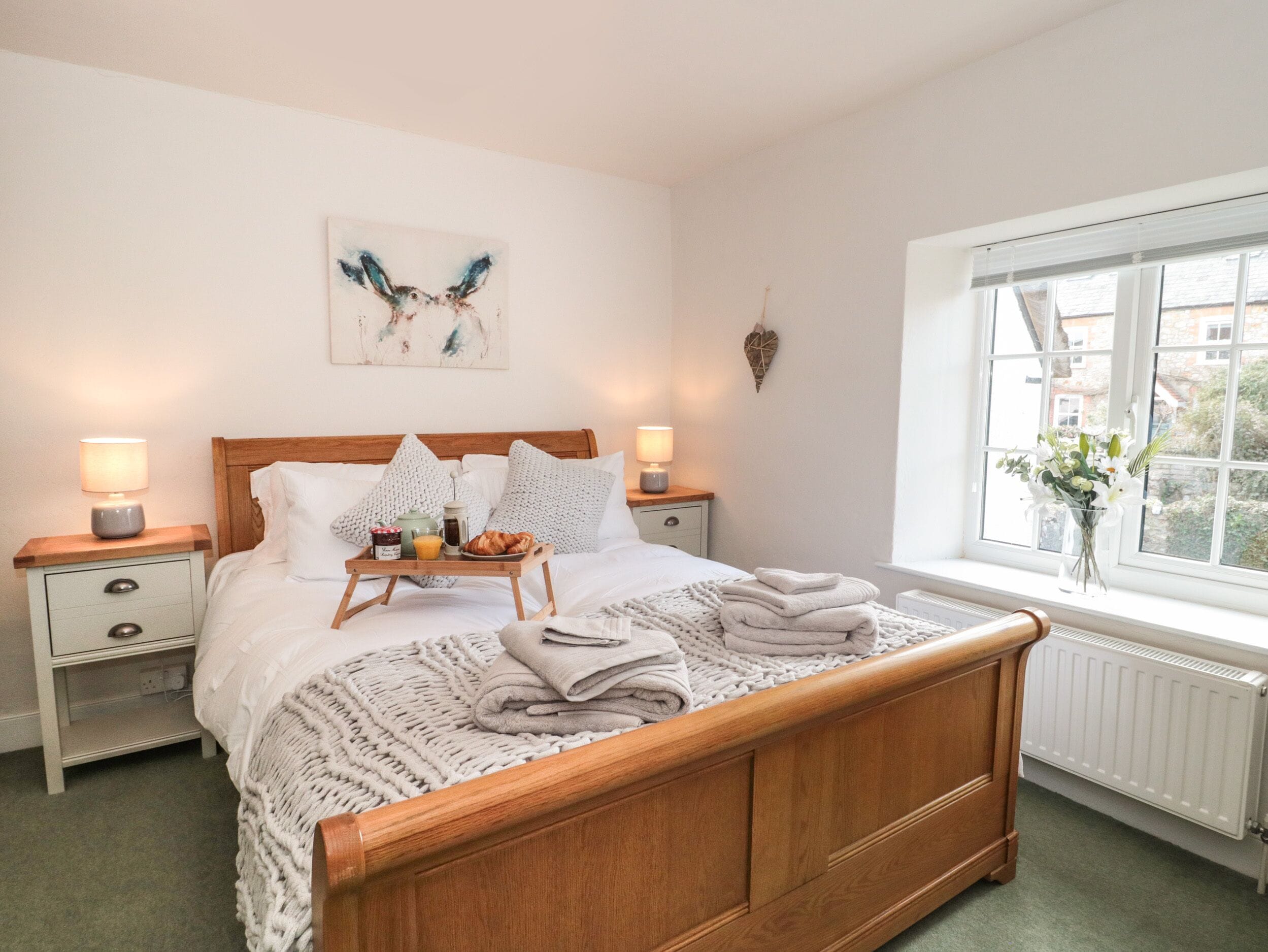 A neatly made bed with a tray of breakfast items, two bedside lamps, a wall art piece, and a window with a view of buildings.