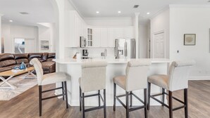 A modern kitchen with white cabinets, a stainless steel refrigerator, and a bar area with bar stools. - Spacious Ocean Retreat~Minutes to the Beach, BBQ, Pool, Beach Gear, and More! (Destin)