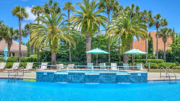 A pool area with lounge chairs and umbrellas, surrounded by palm trees and a building with a tiled roof. - Spacious Ocean Retreat~Minutes to the Beach, BBQ, Pool, Beach Gear, and More! (Destin)