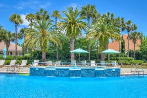 A pool area with lounge chairs and umbrellas, surrounded by palm trees and a building with a tiled roof. - Spacious Ocean Retreat~Minutes to the Beach, BBQ, Pool, Beach Gear, and More! (Destin)