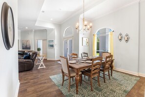 A dining area with a wooden table, chairs, and a chandelier. There is a couch and a painting in the background. - Spacious Ocean Retreat~Minutes to the Beach, BBQ, Pool, Beach Gear, and More! (Destin)