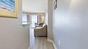 A hallway leading to a living area with a sofa and a dining table, a painting on the wall, and a view of the outdoors.
