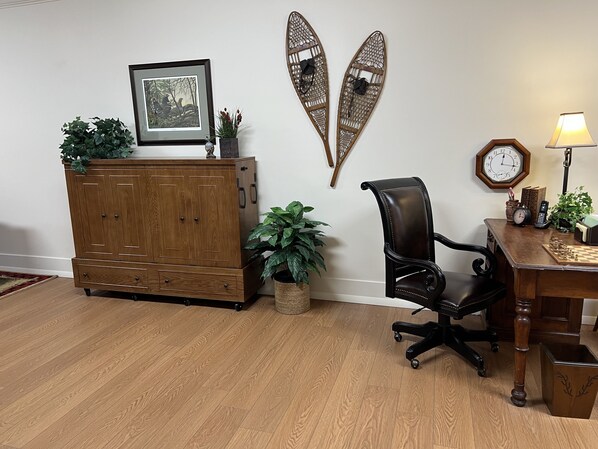 Un mueble de madera con cajones, una silla de oficina negra, un escritorio con lámpara, un reloj y dos elementos decorativos colgados en la pared.