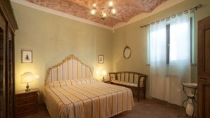 Ein Schlafzimmer mit einem großen Bett, einem Sessel, einem Nachttisch, einem Spiegel und einem Fenster mit Vorhängen.