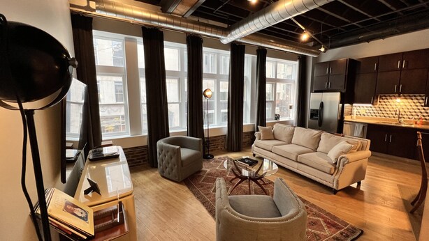 A modern living room with a sofa, armchair, and coffee table. There are large windows with curtains, a lamp, and a kitchen area in the background. - Ambrose Lofts - 1 bedroom overlooking 4th Ave Downtown (Nashville)