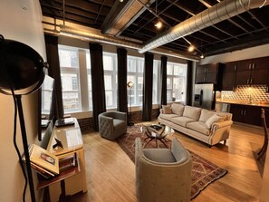 A modern living room with a sofa, armchair, and coffee table. There are large windows with curtains, a lamp, and a kitchen area in the background. - Ambrose Lofts - 1 bedroom overlooking 4th Ave Downtown (Nashville)