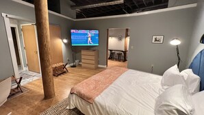 A bedroom with a bed, a desk, a chair, a dresser, a TV, and a lamp. - Ambrose Lofts - 1 bedroom overlooking 4th Ave Downtown (Nashville)