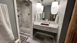 A modern bathroom with a glass-enclosed shower, a marble sink, and a large mirror with decorative lights. - Ambrose Lofts - 1 bedroom overlooking 4th Ave Downtown (Nashville)