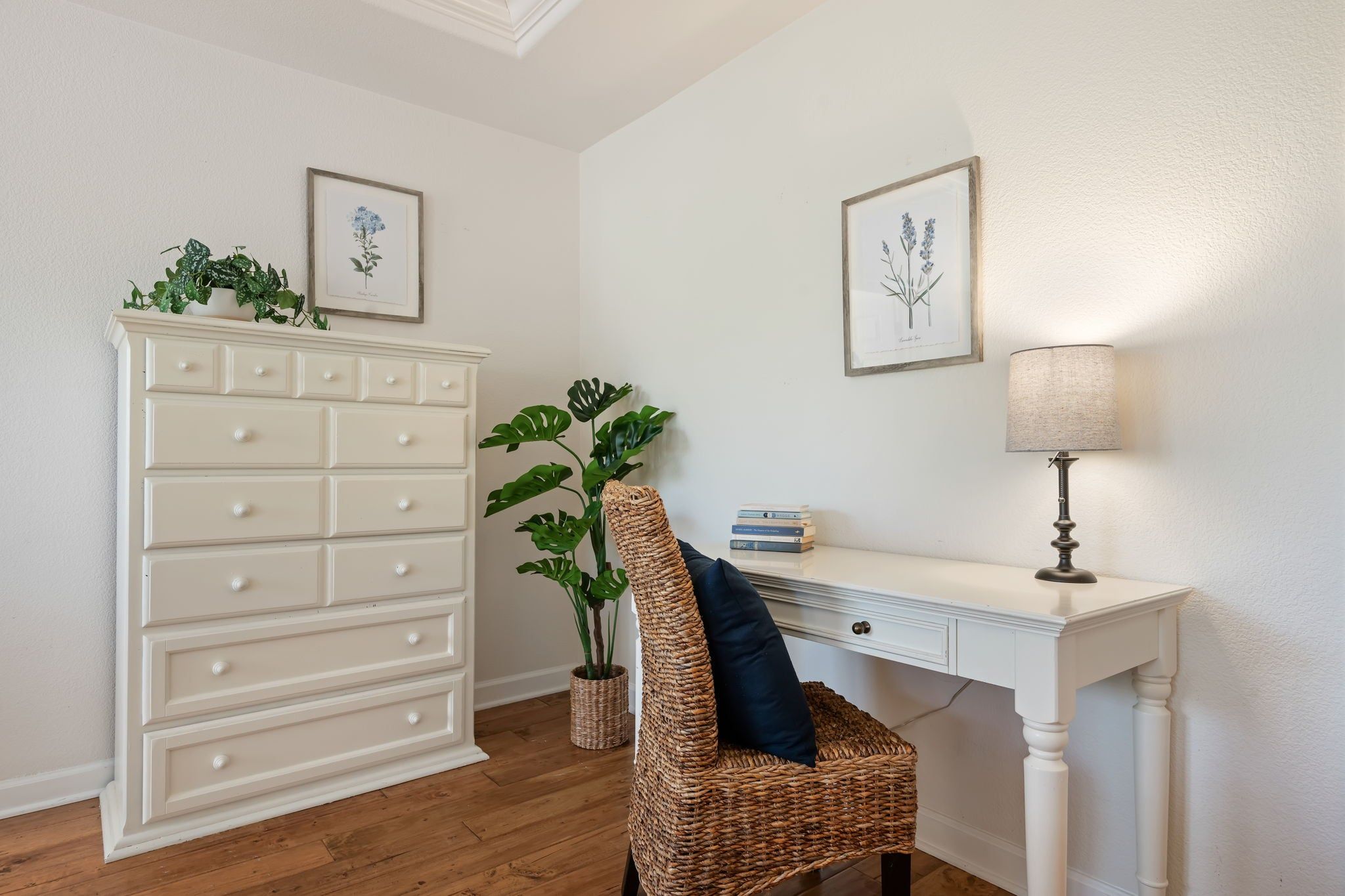 A room with a white dresser, a wicker chair, a desk with a lamp, and framed botanical illustrations on the wall.