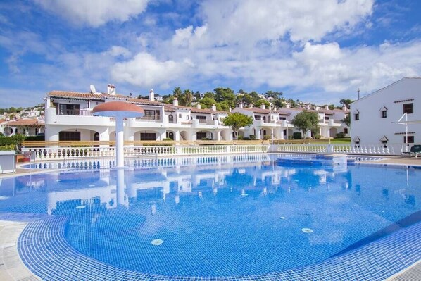 A swimming pool with a tiled deck, surrounded by white buildings with red-tiled roofs, under a clear sky.