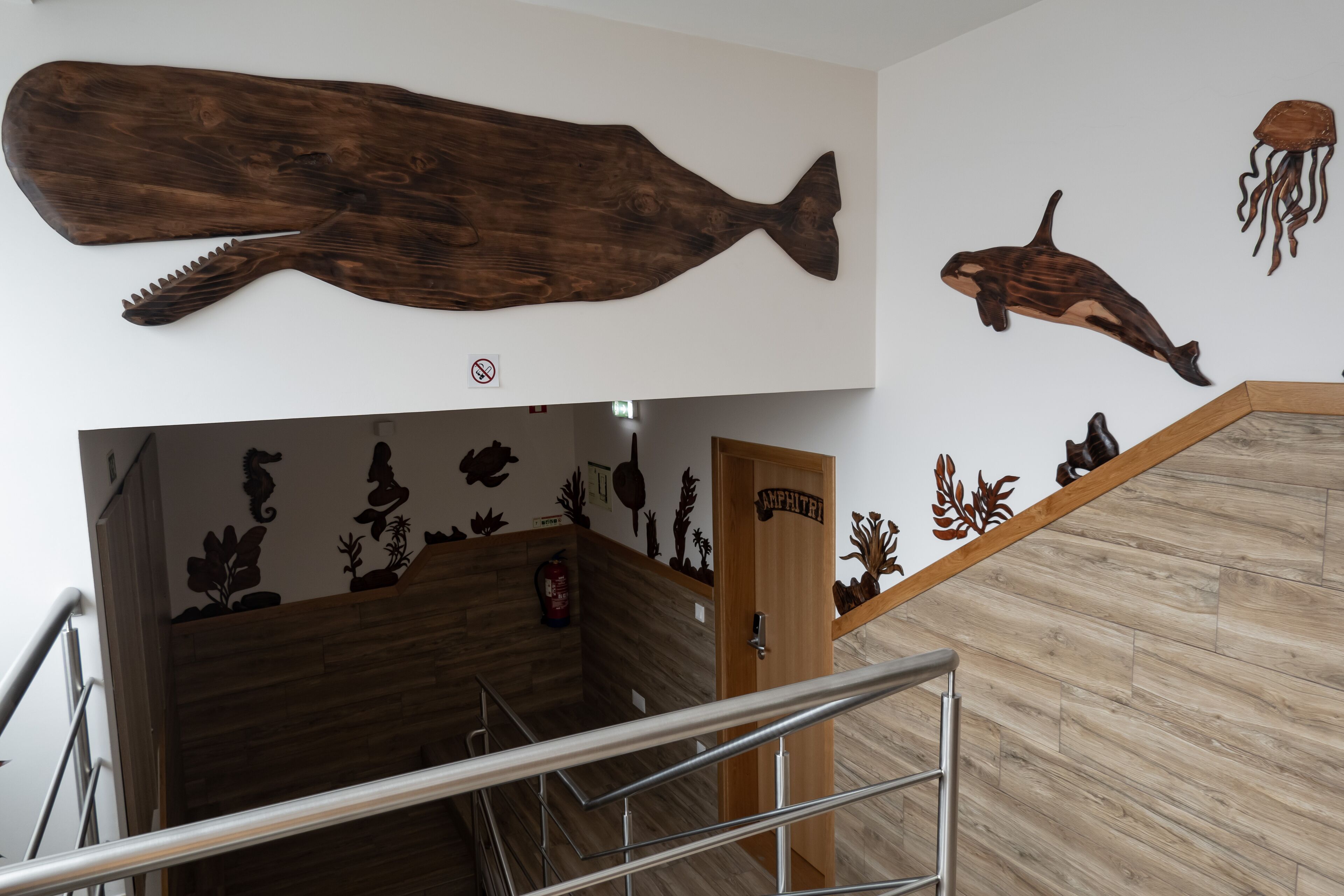 Un escalier avec une rampe en bois, une porte en bois portant l’inscription « AMPHITRIT », et des œuvres d’art murales représentant un poisson, un dauphin et une méduse.
