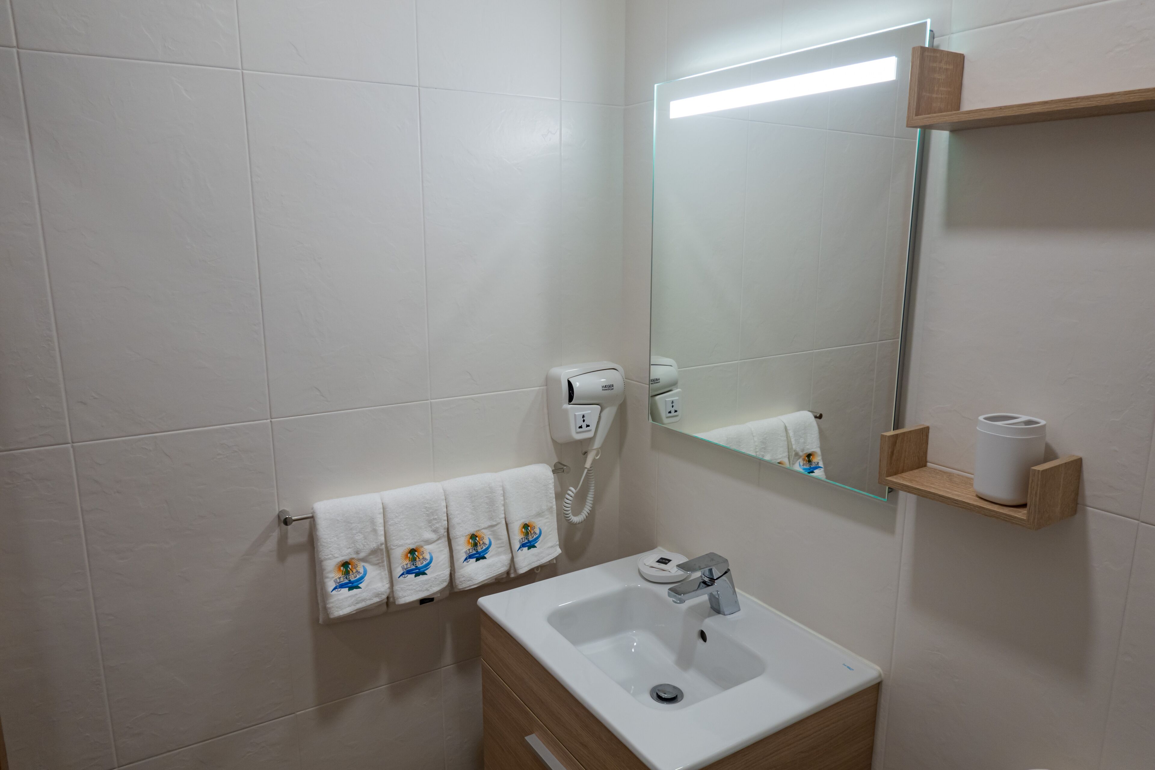 A bathroom with a white sink, a mirror, a towel rack with three towels, a hairdryer, and a shelf with a roll of toilet paper.