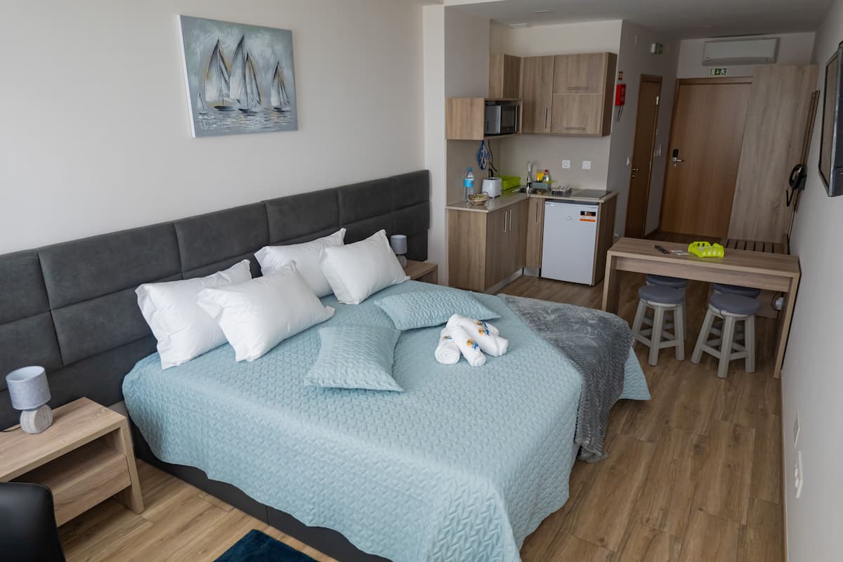 Un dormitorio con cama, almohadas, una mesita de noche, un escritorio con taburetes, una kitchenette y una puerta.