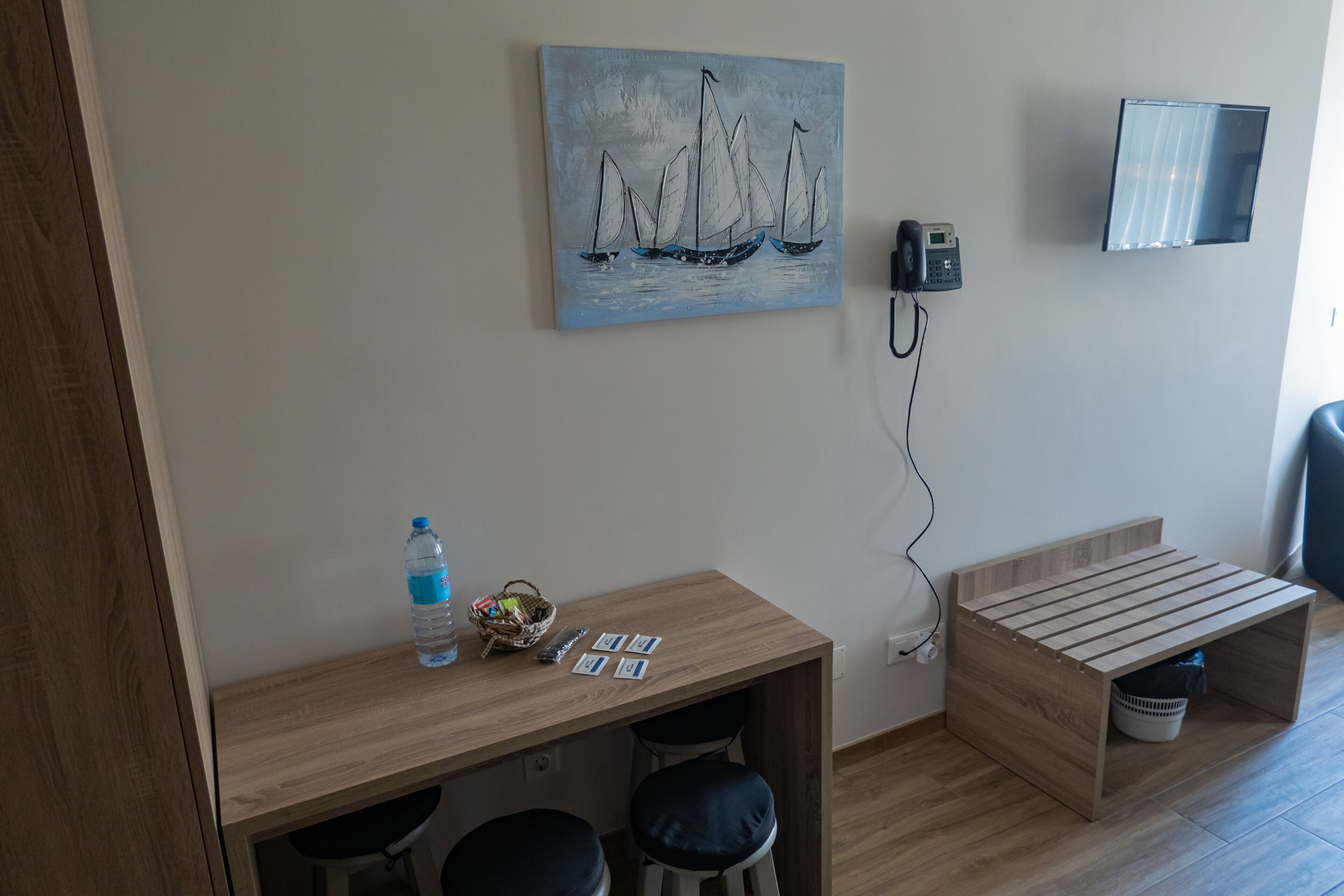 A small room with a wooden desk, two stools, a painting of sailboats, a phone, and a TV mounted on the wall.
