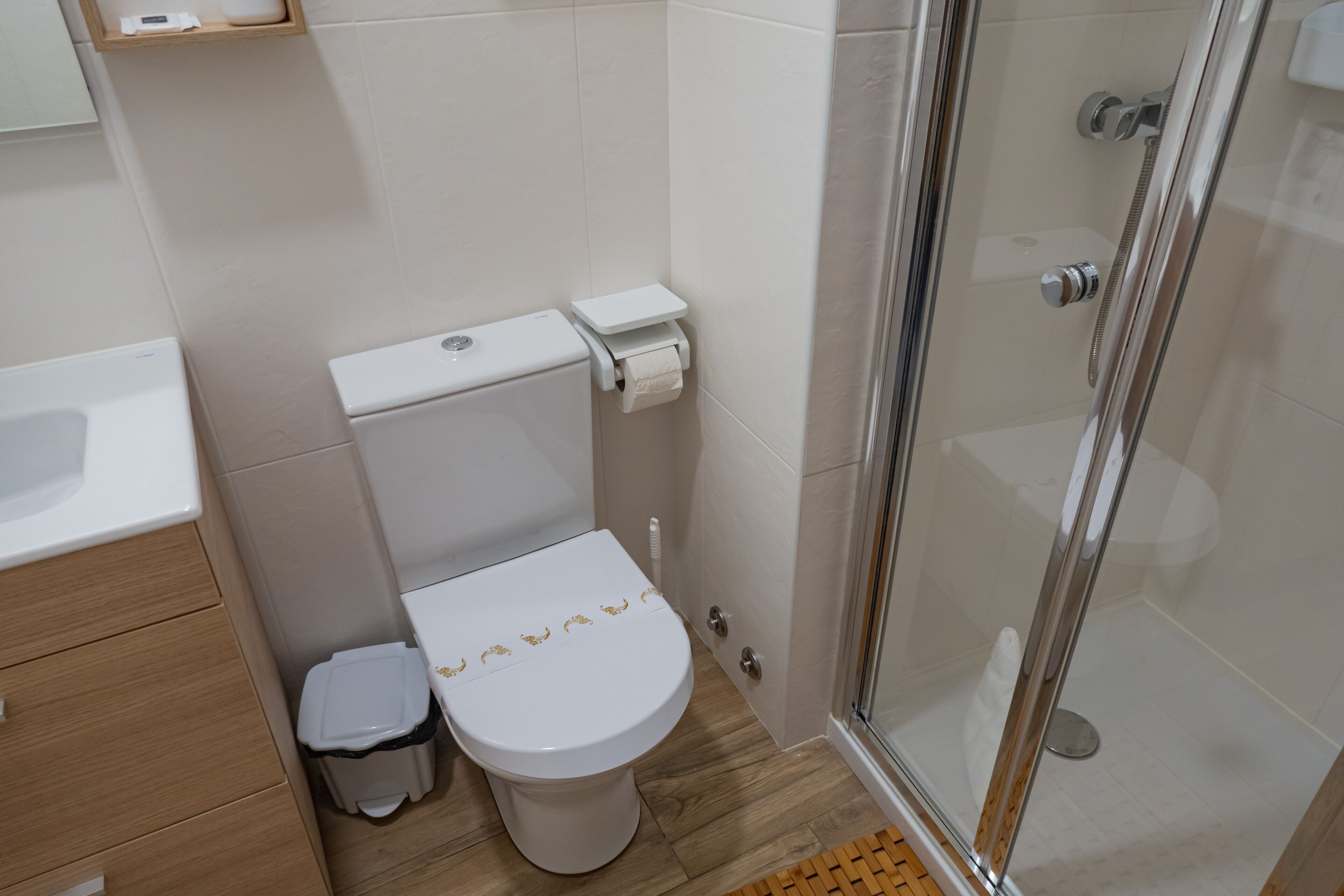 Un baño moderno con inodoro, un cubo de basura pequeño, cabina de ducha de vidrio y suelo de madera.