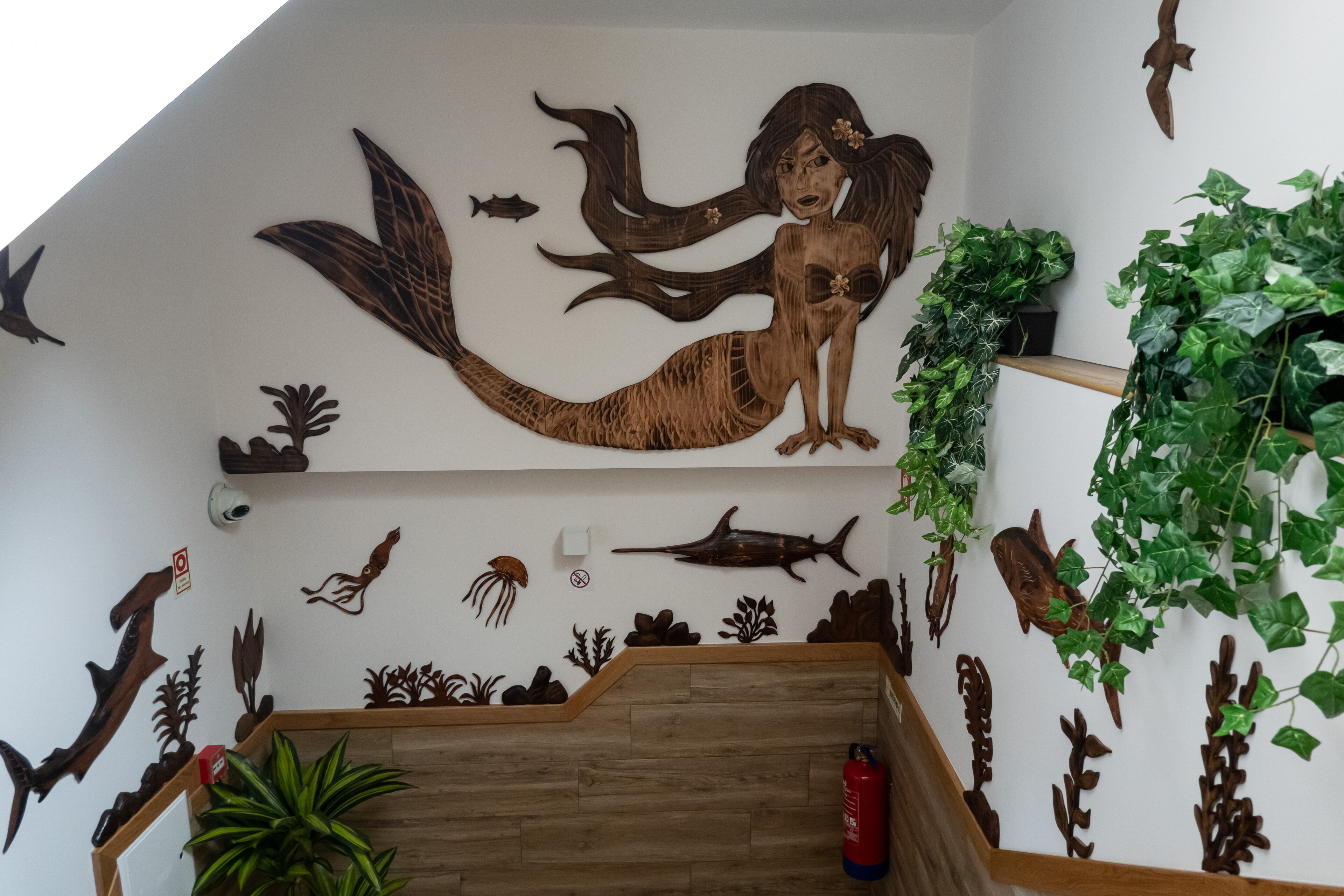 Una escalera con detalles de madera, rodeada de decoraciones marinas fijadas en las paredes, incluyendo una sirena, peces y plantas marinas.