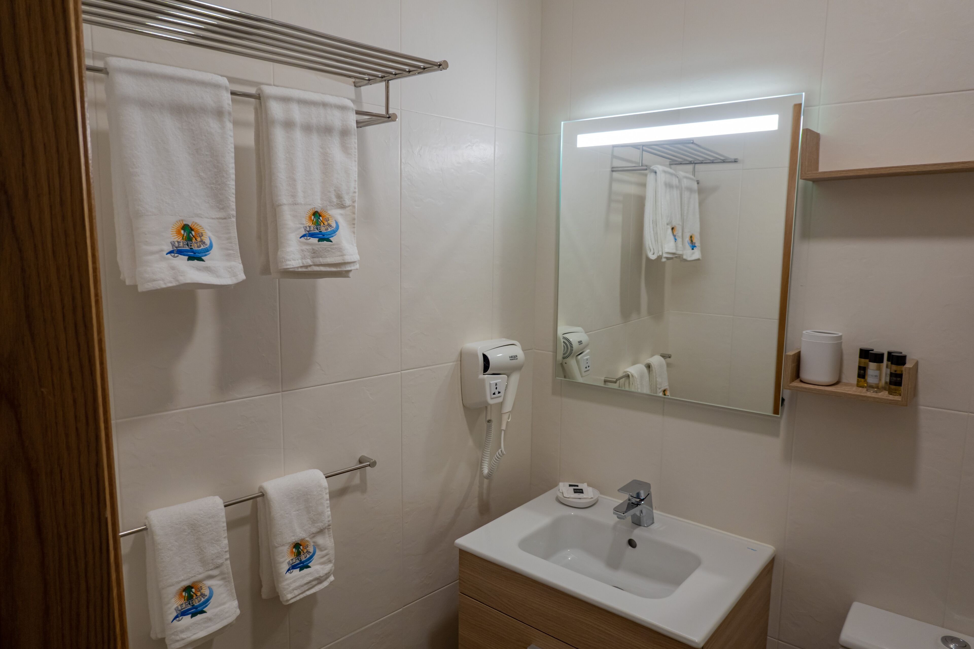 A bathroom with white tiles, a sink, a mirror, and towels with a boat design.