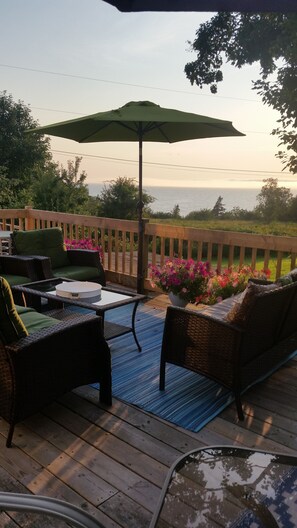 Een houten terras met buitenmeubilair, waaronder stoelen en een tafel, met uitzicht op een tuin met bloemen en een omheining.