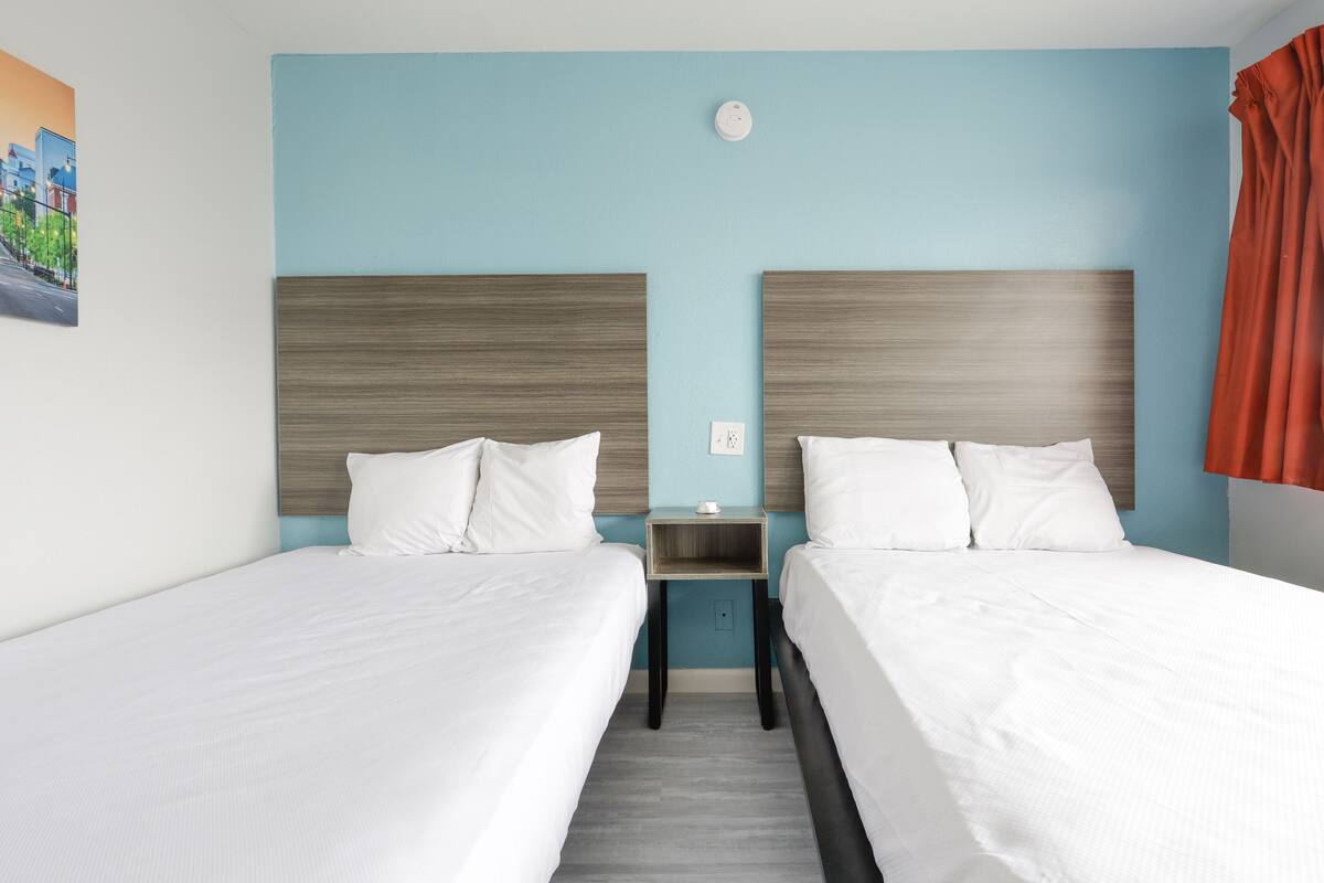 Quarto de hotel com duas camas, uma mesa de cabeceira, uma luminária na parede e um quadro pendurado.