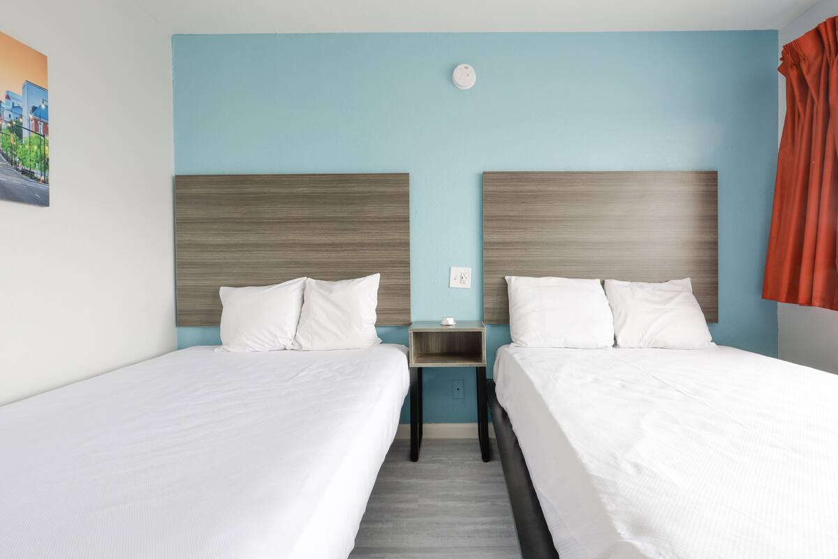 Quarto de hotel com duas camas, uma mesinha de cabeceira e um candeeiro fixado na parede.