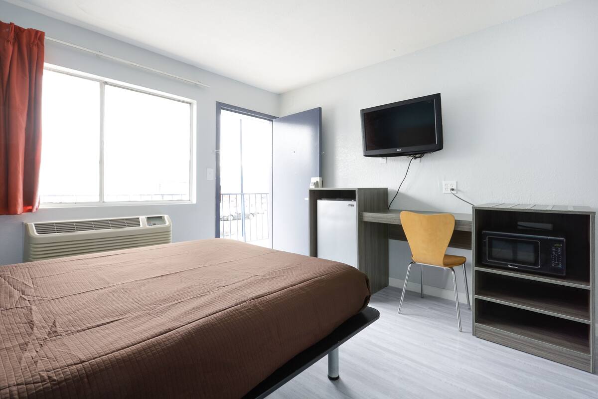Quarto de hotel com cama, escrivaninha, cadeira, televisão e micro-ondas.