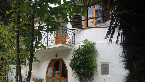 Ein weißes Gebäude mit einer Holztür und einem Balkon. Pflanzen und Steine sind am Eingang angeordnet.