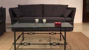 Ein Wohnzimmer mit einem dunkelgrauen Sofa, einem Glastisch mit zwei Kerzen und drei Landschaftsbildern an der Wand.