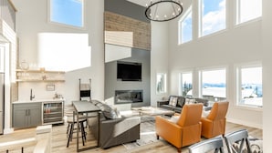 A spacious living area with a fireplace, a TV mounted on the wall, and a large window offering a view of the outdoors.