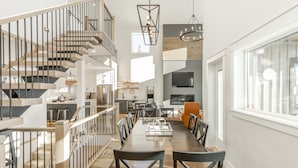 A modern kitchen with a central island, a dining area with a long table and chairs, and a staircase with a railing.